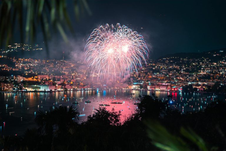 Fireworks illuminate the night sky over Lake Lugano, showcasing a vibrant cityscape reflection.